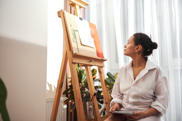 Femme peintre en chemise blanche debout devant le chevalet pensant et regardant la toile