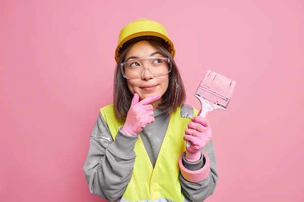 femme peintre garde la main sur le menton considère que la réparation de la maison porte des lunettes de protection casque et uniforme tient des poses de pinceau