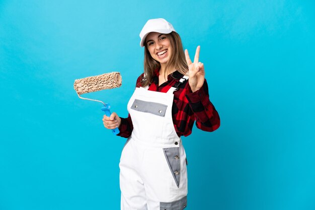 Femme peintre sur mur bleu isolé souriant et montrant le signe de la victoire