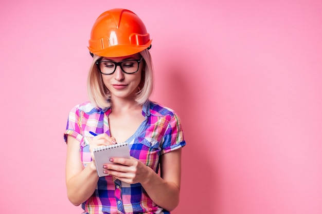 Femme peintre utilisant un rouleau pour remettre à neuf la couleur du mur rose à l'intérieur. Femme souriante en chemise à carreaux d'affaires portant un casque de constructeur peignant les murs de l'appartement.