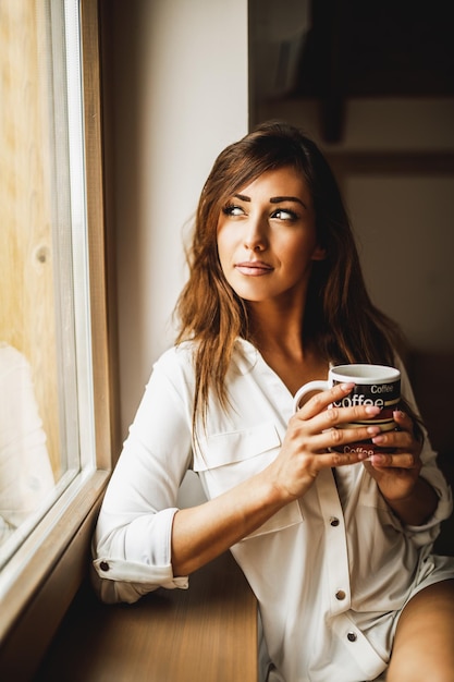 Une femme pensive boit du café le matin et regarde par la fenêtre.