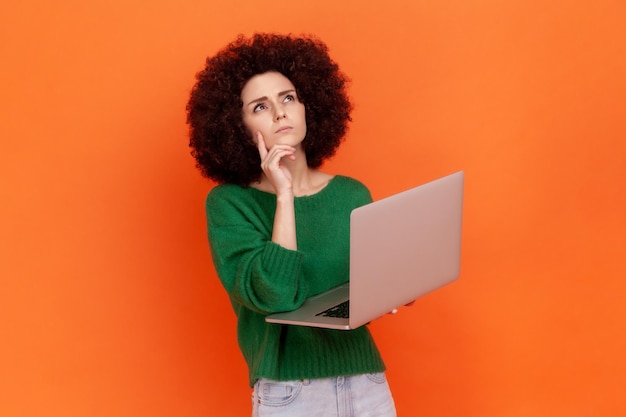 Femme pensive avec une coiffure Afro portant un pull vert de style décontracté travaillant sur un ordinateur portable, tenant le menton, pensant à un nouveau projet, levant les yeux. Studio intérieur tourné isolé sur fond orange.