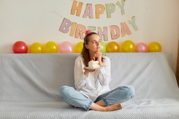 Femme pensive portant une chemise blanche et un jean assis sur un canapé avec les jambes croisées, tenant un gâteau avec une bougie, regardant de côté et faisant un vœu, célébrant l'anniversaire, décoration festive sur fond.