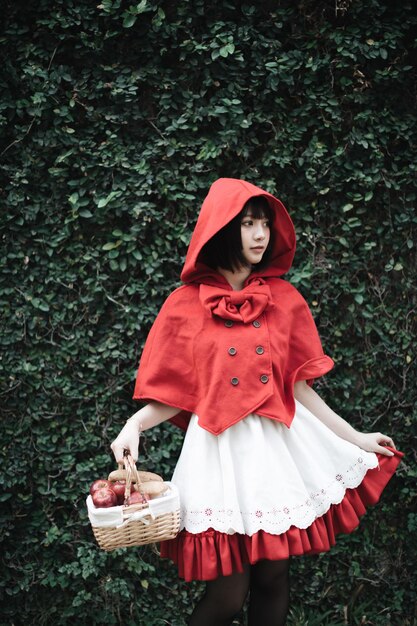 Photo femme en petit costume de capuche rouge tenant un panier alors qu'elle se tient contre les plantes