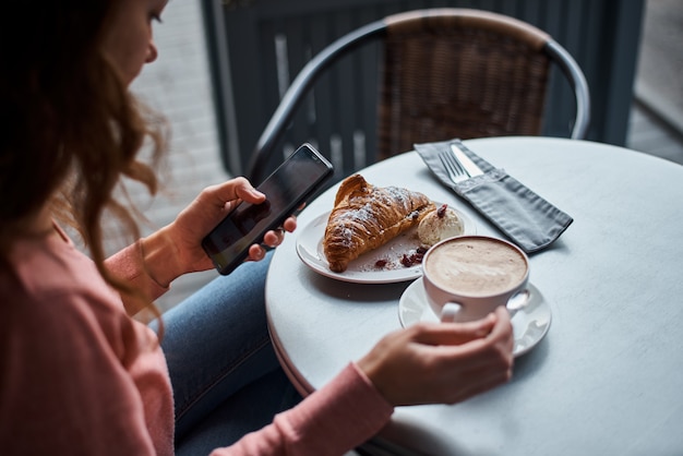 Femme petit déjeuner au café et à l'aide de smartphone. Fille discutant et utilisant Internet avec téléphone pendant la pause-café avec un croissant