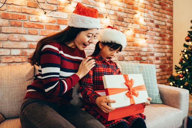 Femme et petite fille ouvrent curieusement une grande boîte cadeau rouge le lendemain de Noël. famille portant des chapeaux de père Noël célébrant Noël dans une chambre confortable et décorée à la maison. concept de cadeau de noël surpris.