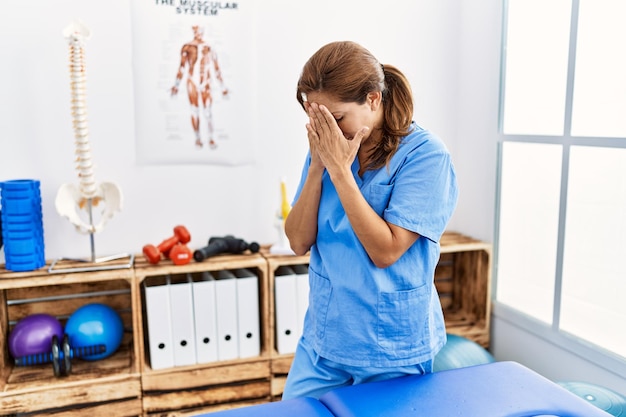 Femme physiothérapeute hispanique d'âge moyen travaillant à la clinique de récupération de la douleur avec une expression triste couvrant le visage avec les mains tout en pleurant le concept de dépression