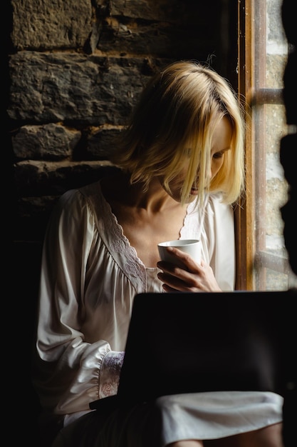 Femme pigiste avec une tasse de café et un ordinateur portable près de la fenêtre Femme d'âge moyen dans une chemise blanche