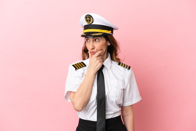 Femme pilote d'âge moyen d'avion isolée sur fond rose ayant des doutes et avec une expression de visage confuse