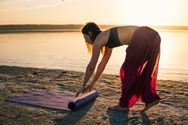 Une femme sur la plage au coucher du soleil commence une formation de yoga avec un tapis de yoga. Entraînement d'échauffement naturel du matin