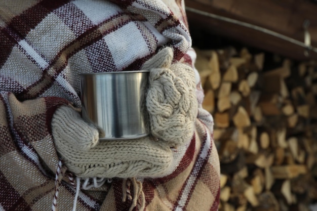 Une femme en plaid tient une tasse en métal sur fond de bois de chauffage