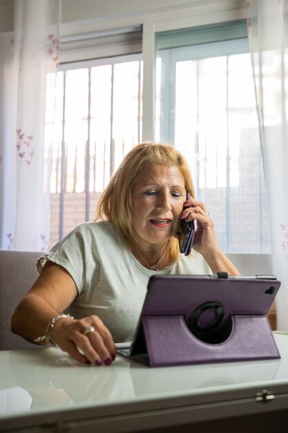 Une femme plus âgée assise à la maison tout en parlant sur son téléphone portable avec quelqu'un d'autre et se divertit avec sa tablette