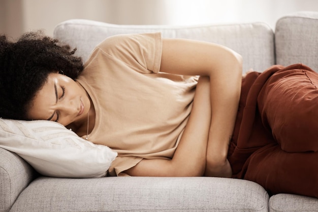 Femme Pms et maux d'estomac sur un canapé de la période menstruelle et crampes dans un salon Mal au ventre femme noire et douleur due à l'endométriose problème de digestion ou de constipation sur un canapé à la maison