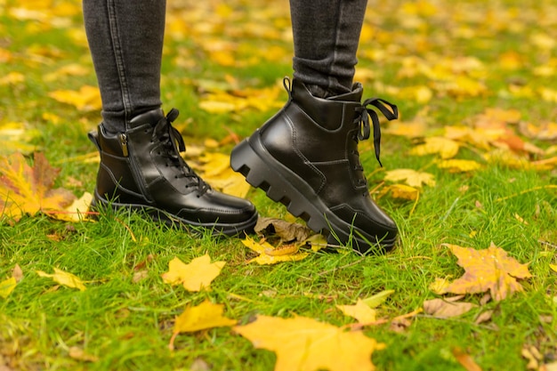 Une femme portant des bottes noires se tient dans l'herbe avec des feuilles jaunes sur le sol.