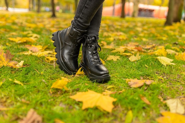 Une femme portant des bottes noires se tient dans l'herbe avec des feuilles jaunes sur le sol.