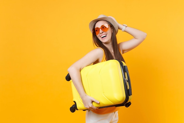 une femme portant un chapeau et des lunettes de soleil tient une valise jaune