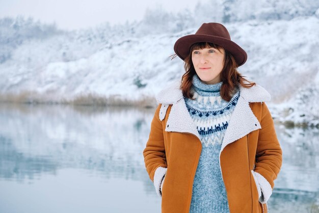 Femme portant un chapeau et une veste chaude debout sur les rives d'un lac et d'une forêt enneigés