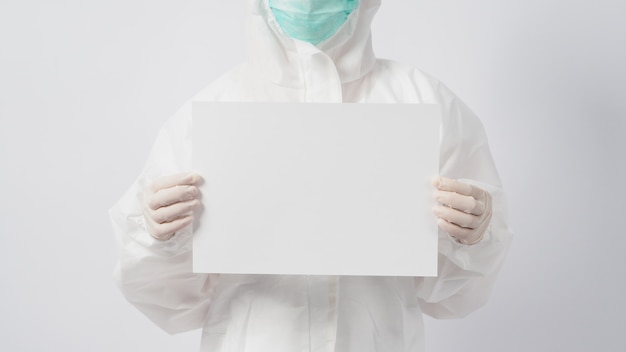 Une femme portant un EPI et un masque facial et une main avec des gants tient du papier A4 vierge sur fond blanc.
