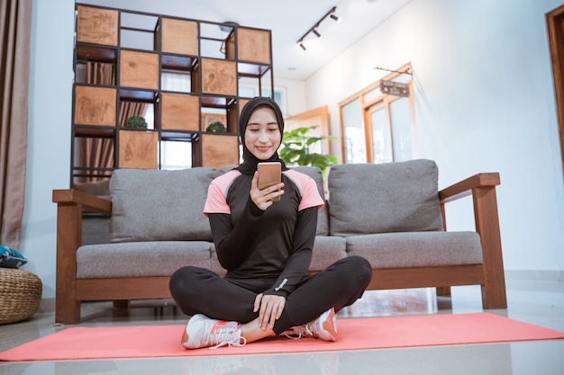 Une femme portant un hijab sportswear est assise les jambes croisées sur un tapis tout en utilisant son téléphone portable avec le mur du canapé