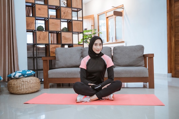 Une femme portant un hijab sportswear est assise sur le sol sur un tapis de yoga dans le salon