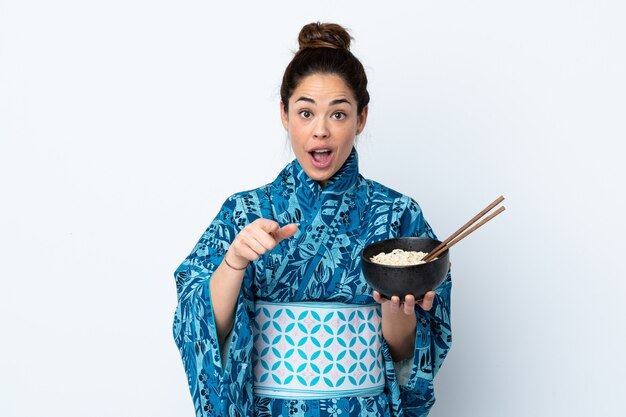 Femme portant un kimono sur blanc isolé surpris et pointant vers l'avant tout en tenant un bol de nouilles avec des baguettes