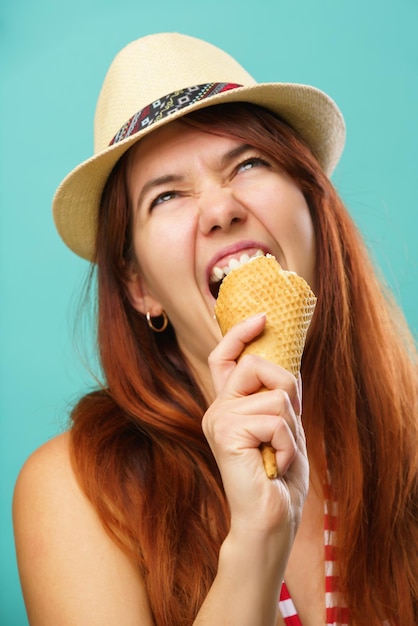 Une femme portant un maillot de bain et un chapeau de paille mange de la glace à partir d'une tasse sur fond turquoise coloré