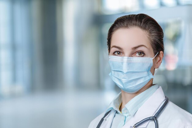 Une femme portant un masque et un manteau blanc se tient devant un arrière-plan flou.