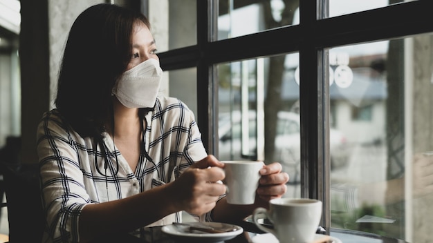 Une femme portant un masque médical est assise dans un café et regarde par la fenêtre avec des yeux tristes, elle en a marre de l'épidémie de virus et veut vivre une vie normale.