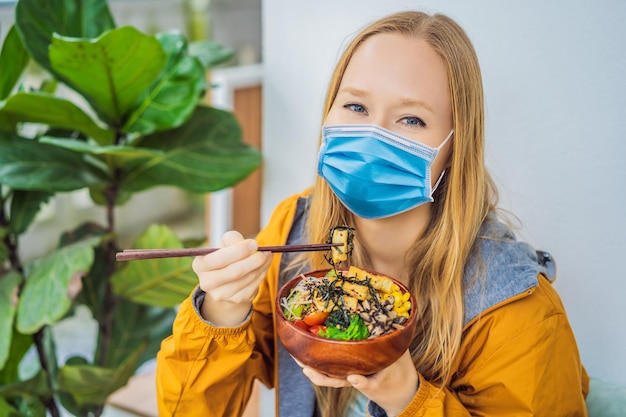 Femme portant un masque médical pendant le coronavirus covid mangeant un bol de poke bio cru avec du riz et