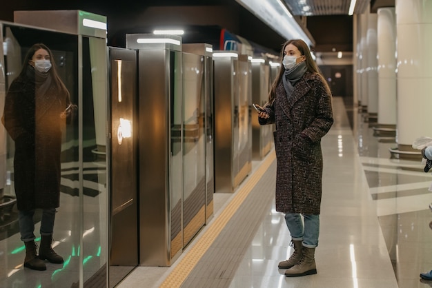 Une femme portant un masque médical pour éviter la propagation du coronavirus cache un smartphone en attendant un train sur le quai du métro. Une fille portant un masque chirurgical garde une distance sociale.