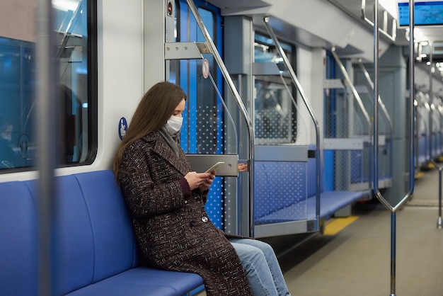 Une femme portant un masque médical pour éviter la propagation du coronavirus est assise et utilise un smartphone dans une voiture de métro moderne