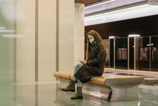 Une Femme Portant Un Masque Médical Pour éviter La Propagation Du Coronavirus Est Assise Et Utilise Un Smartphone Sur Le Quai Du Métro. Une Fille Portant Un Masque Chirurgical Garde Une Distance Sociale Dans Le Métro.