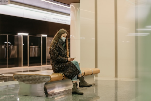 Une femme portant un masque médical pour éviter la propagation du coronavirus est assise et utilise un smartphone sur le quai du métro. Une fille portant un masque chirurgical garde une distance sociale dans le métro.