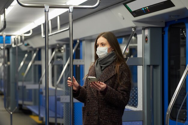 Une femme portant un masque médical pour éviter la propagation du coronavirus est debout et tient un téléphone dans une voiture de métro moderne