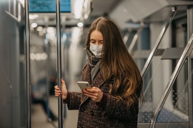 Une femme portant un masque médical pour éviter la propagation du coronavirus est debout et utilise un smartphone dans une voiture de métro moderne. Une fille portant un masque chirurgical fait défiler les nouvelles sur son téléphone portable dans un train.