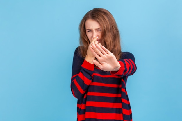 Femme portant un pull rayé saisissant le nez, grimaçant de dégoût et montrant un geste d'arrêt, exprimant une répulsion pour la puanteur, les gaz de pet, l'odeur intolérable. Studio intérieur tourné isolé sur fond bleu.