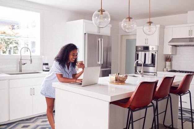 Femme portant un pyjama debout dans la cuisine travaillant à domicile sur un ordinateur portable