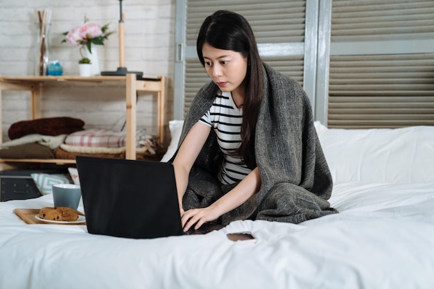 femme portant un t-shirt rayé et assis sur un lit blanc tapant clavier ordinateur portable avec plateau de petit déjeuner pain et tasse de thé chaud. femme asiatique travaillant sur un ordinateur portable et savourant un café le matin