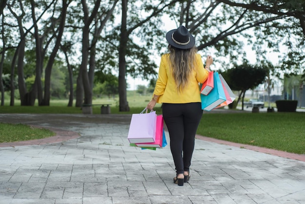 Femme portant une veste jaune marchant dans un jardin avec des sacs à provisions multicolores Shopping concept