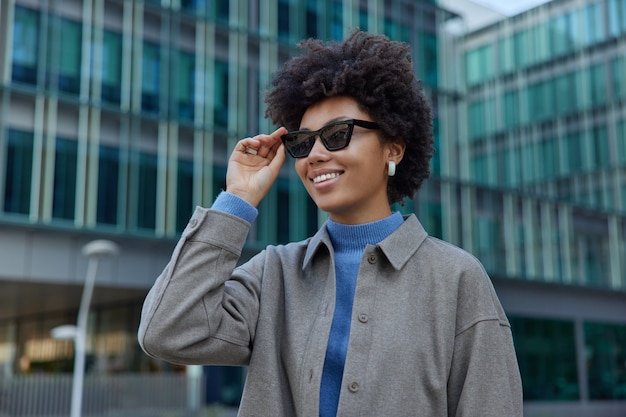 une femme porte des lunettes de soleil à la mode à col roulé et un manteau se promène en ville contre des bâtiments modernes admire une belle vue se sent heureuse profite de son temps libre