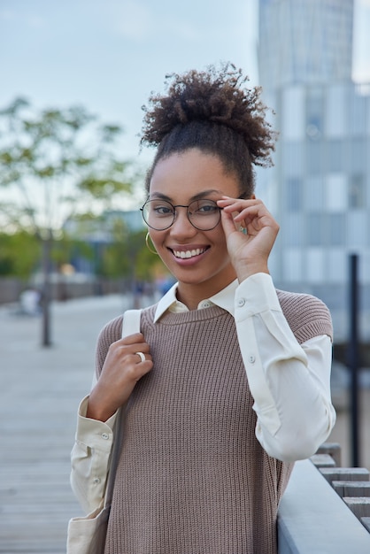 la femme porte des lunettes transparentes et des vêtements soignés sourit joyeusement porte un sac en tissu se promène en ville seule profite du beau temps pendant le week-end