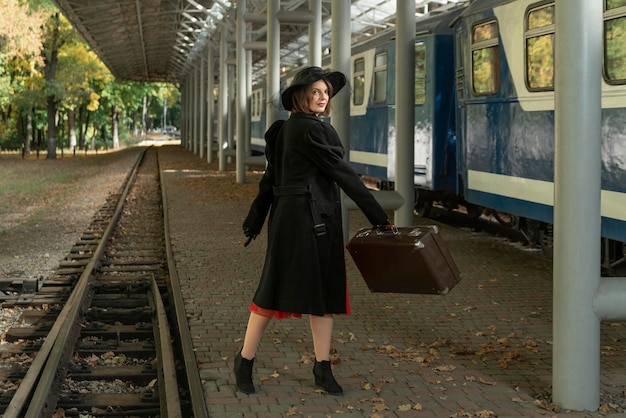 Femme porte des vêtements d'un manteau noir des années 40 et un chapeau tient une valise rétro Fille sur la plate-forme près du train