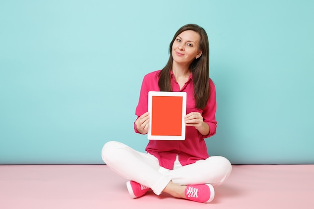 Femme portrait pleine longueur en chemise rose chemisier, pantalon blanc assis sur le sol tenir tablet pc isolé sur mur pastel bleu rose vif.