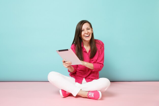 Femme portrait pleine longueur en chemise rose chemisier, pantalon blanc assis sur le sol tenir tablet pc isolé sur mur pastel bleu rose vif.