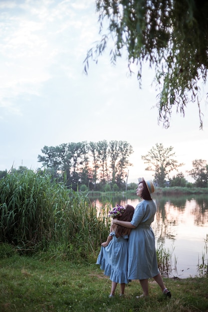 Photo femme posant en plein air dans la nature avec bouquet de fleurs et profitez de la vue magnifique sur le lac et le soleil. la mère étreignant la fille ou la fille. des mannequins vêtus de robes bleues. bonne fête des mères