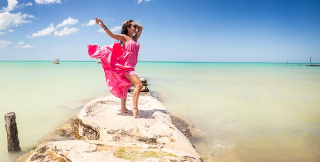 Femme posant en robe rose se dresse sur un rocher à côté d'une mer exotique dans une destination de vacances d'été