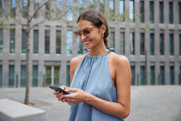 une femme pose en milieu urbain pendant une journée ensoleillée porte des lunettes de soleil robe bleue utilise un gadget pour smartphone pour les discussions en ligne utilise un navigateur pour trouver un itinéraire