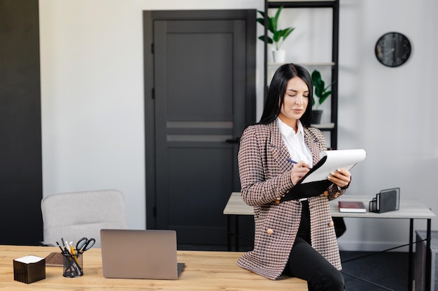 Une femme positive assise à une table de bureau écrivant des notes sur papier tout en travaillant sur un projet dans un lieu de travail moderne