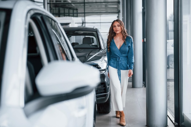 Une femme positive en chemise bleue marche près d'une voiture neuve dans un salon de l'auto