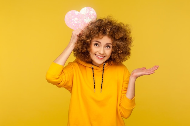 Femme positive avec une coiffure afro et un sourire charmant tenant un coeur de papier rose sur la tête et écartant la paume portant un sweat à capuche de style décontracté Prise de vue en studio intérieure isolée sur fond jaune